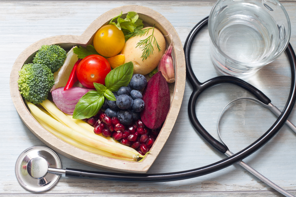 photo from above of a heart-shaped bowl filled with fruits and vegetables and a glass of clear liquid and a stethoscope arranged next to it