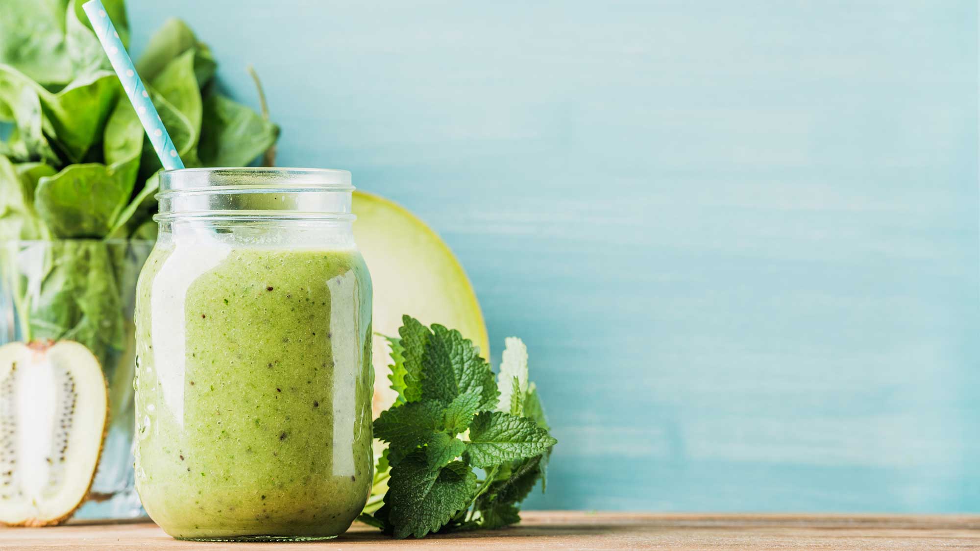 image of a green smoothie in a mason-style jar with fresh fruit and leafy greens in the background
